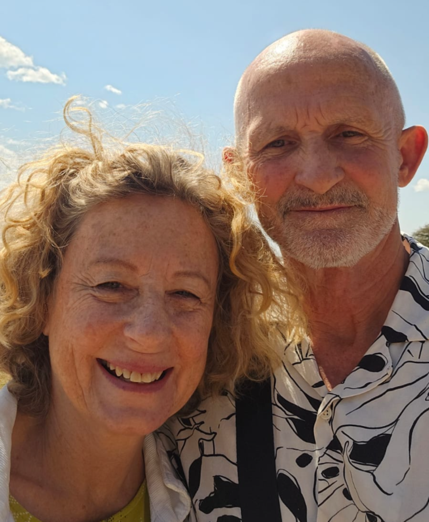 A woman and a man with light hair and beards pose outdoors in bright sunlight, both smiling at the camera against a clear blue sky.