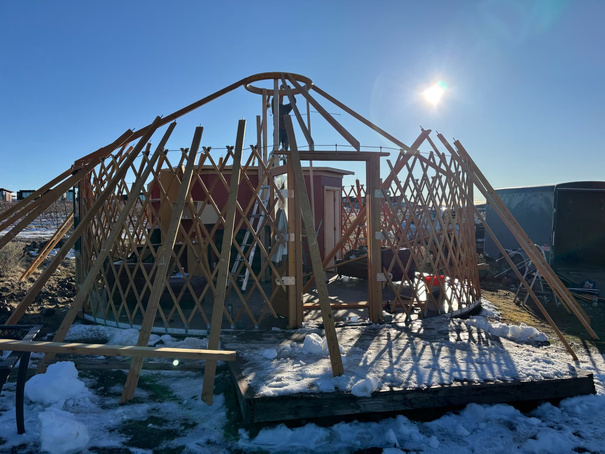 A partially constructed yurt with exposed wooden framework sits on a snowy ground under a clear sky with the sun shining.