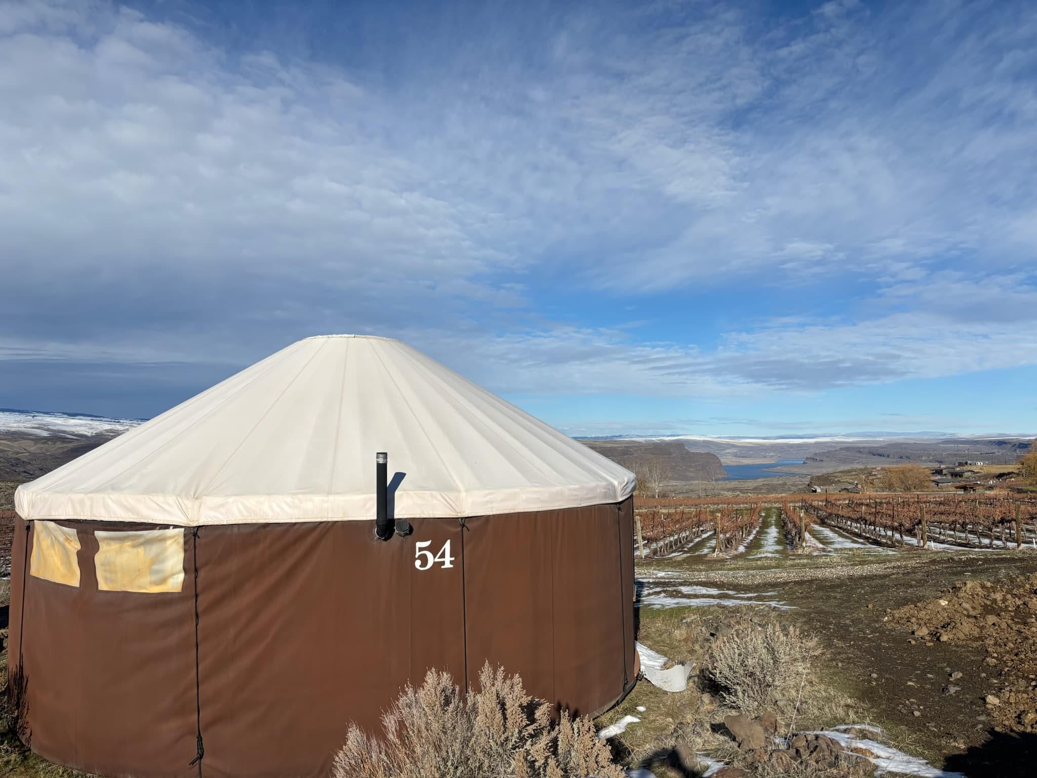 A round brown yurt labeled "54" stands in front of a vineyard and hilly landscape under a partly cloudy sky.