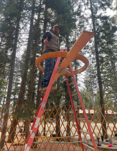 A man stands on a wooden plank balanced between a ladder and a circular frame, with trees and a wooden lattice in the background.