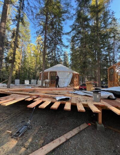 A person stands in front of a partially constructed yurt on a wooden platform surrounded by tall pine trees and building materials.