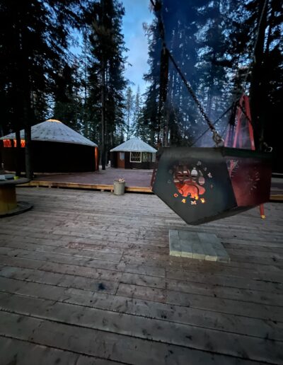 A metal fire pit with a bear paw design hangs over a fire on a wooden deck, with two lit yurts and tall trees in the background at dusk.