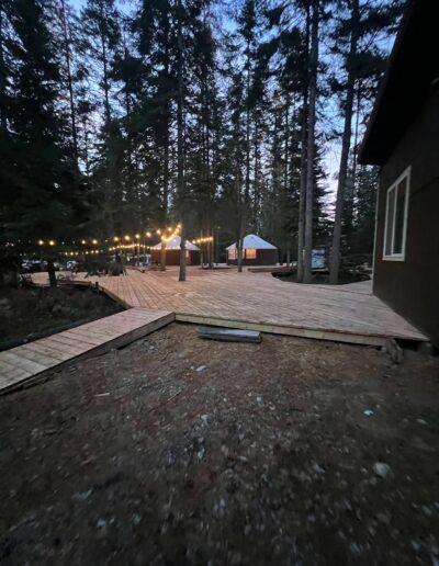 A wooden deck with string lights among tall trees at dusk; a yurt and small structure are visible in the background.