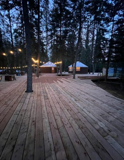 A large wooden deck with string lights and two yurts in a forested area at dusk.