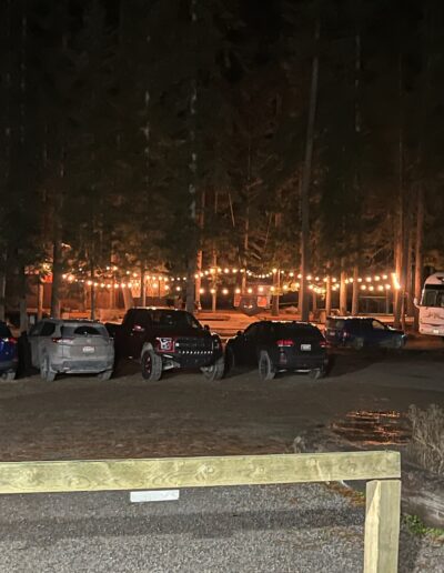 Several cars are parked on a dirt lot at night with string lights illuminating a building and trees in the background.