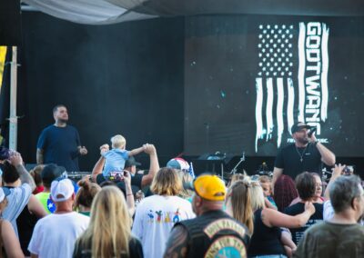 A crowd watches two performers on an outdoor stage with a screen behind them displaying a stylized U.S. flag and the word "GODTWA".