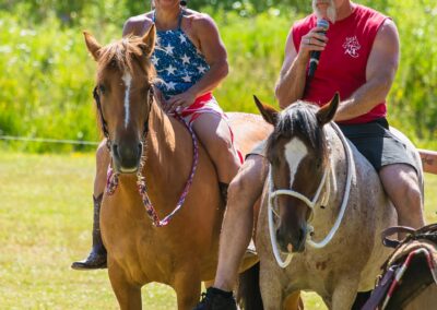 Two adults sit on horses outdoors; the woman smiles, wearing a star-spangled top, while the man in a red tank top drinks from a bottle. Trees and grass are visible in the background.