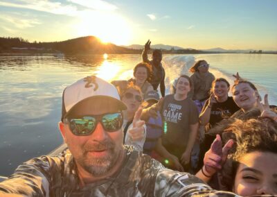 A group of people smile and pose for a selfie on a boat at sunset, with calm water and distant hills in the background.