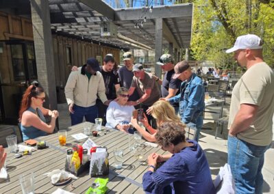 A group of people gather around an outdoor table at a restaurant, with some standing and others sitting, while one person shows something on a phone.