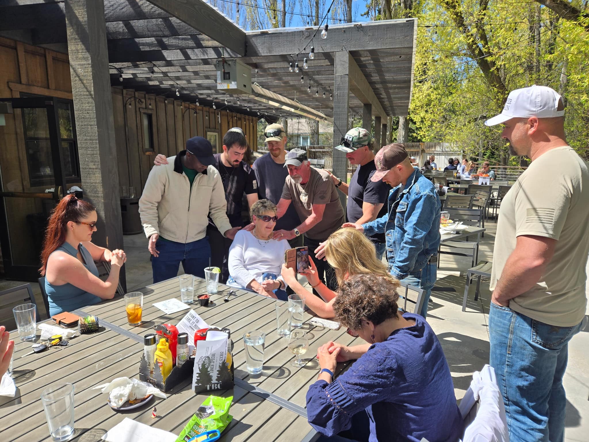 A group of people gather around an outdoor table at a restaurant, with some standing and others sitting, while one person shows something on a phone.