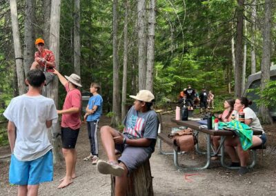 A group of people at a forest campsite; some stand or sit near a picnic table, while others chat by a campfire in the foreground.