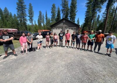 A group of people stand in a line outdoors on gravel in front of a garage and trees under a clear blue sky.