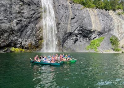 A group of people in colorful canoes paddle on a green lake near a tall waterfall with rocky cliffs and trees in the background.