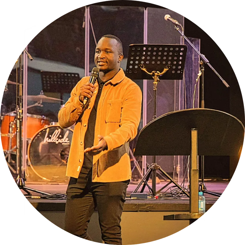 A man speaks into a microphone on stage, standing near a podium with music stands and a drum set visible in the background.