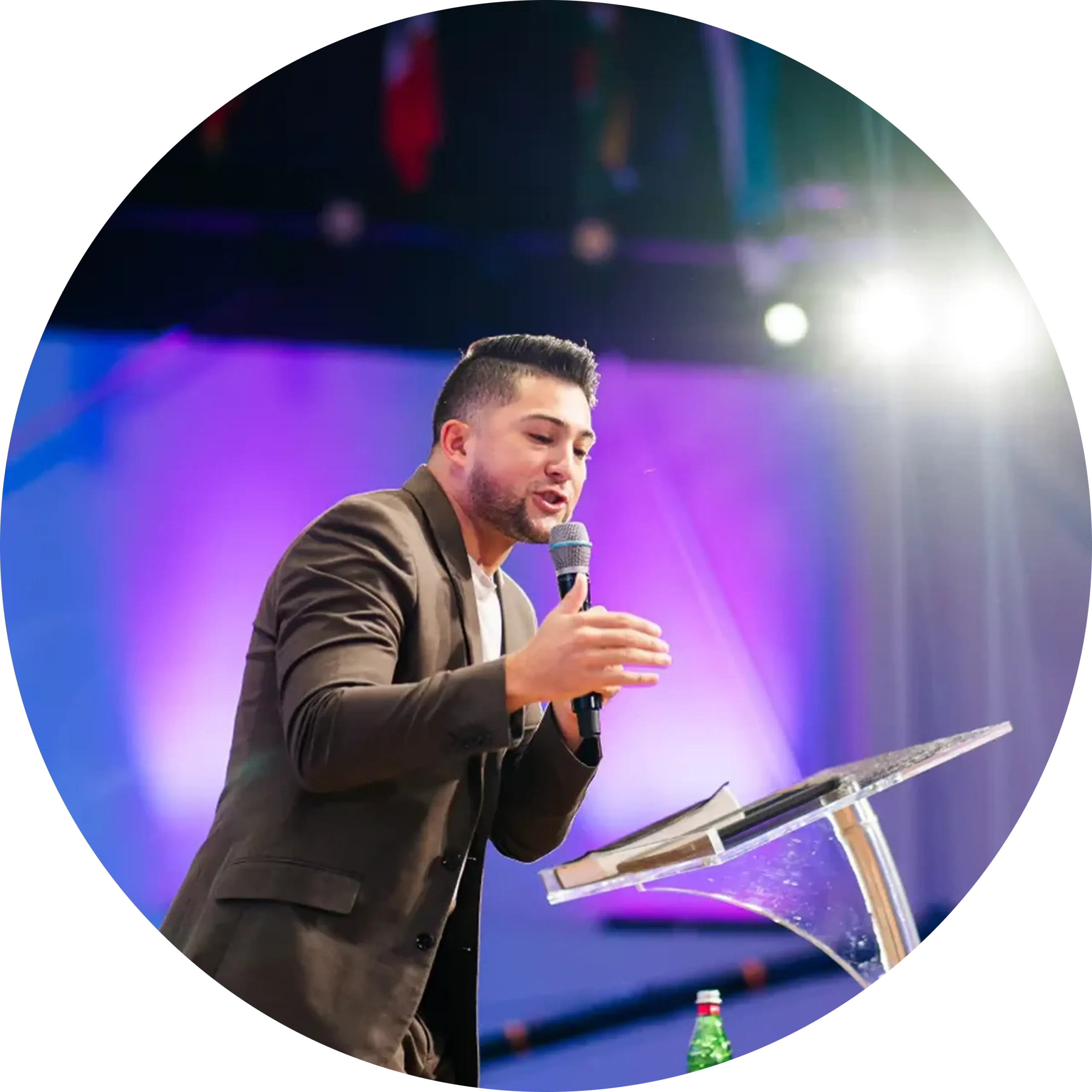A man in a suit speaks into a microphone at a clear podium with an open book and a water bottle, under bright stage lights and a purple-lit background.